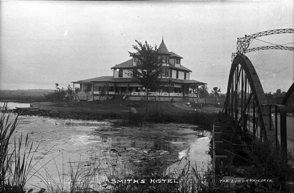 A bridge leads across a body of water to the three-story Smith's (Resort) Hotel. Several people stand on the building's wrap-around porch and the roof features a turret and window dormers.  "Smith's Hotel" and "The Long & Taylor Co." are written on the bottom of the image.