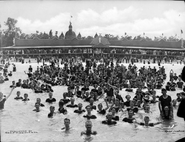 A large group of men, women, and children stand in the water wearing bathing suits while others look on from a pavilion in the background. Several flags can also be seen atop the pavilion, and trees stand in the background.