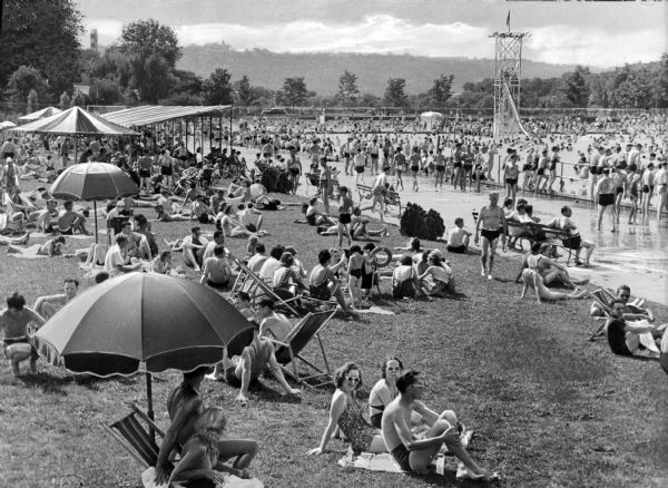 People sunbathe on a crowded beach along the Ohio River.  A shaded pavilion stands on the beach at left and an American flag flies from a slide in the water.