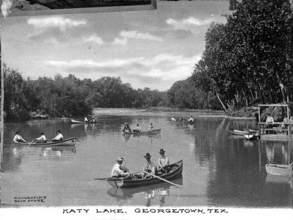 Men paddle rowboats near a dock on Katy Lake. The view features five rowboats in plain view, and several other boats off to the far right. The lake is bordered by trees.