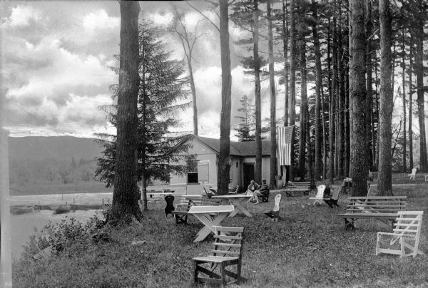 A man and woman sit in front of a cabin in Gibson's Grove on Lake Buell.  Multiple benches and chairs stand on the lawn looking over the water.