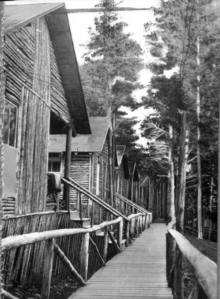 The Cobbossee Colony (Camp?) Boardwalk leads past multiple log cottages in a wooded area.