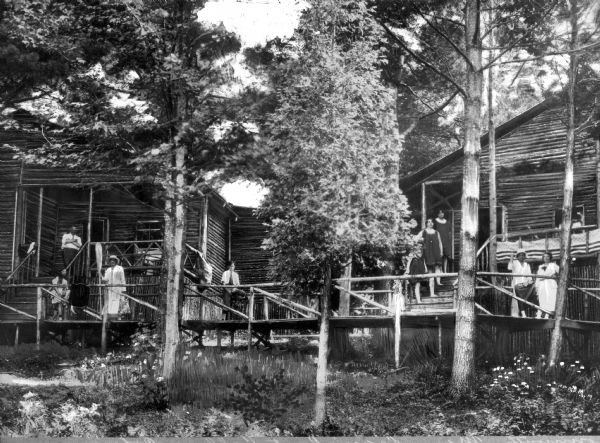 People stand along a log boardwalk in front of log cottages. Trees and foliage surround the buildings.