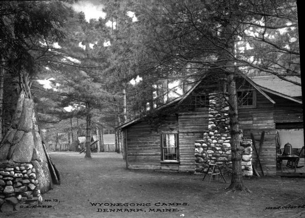 A wooden cabin with a stone chimney stands at Wyonegonic Camp, the oldest girls' camp in the United States. The cabin is surrounded by trees and a tipi stands to the left.