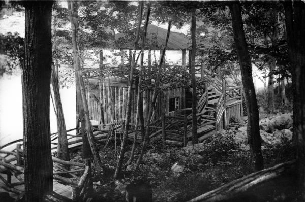 Elevated view of White Birch Lodge.  A log walkway leads to the rustic summer house surrounded by trees.  A body of water stands in the background.