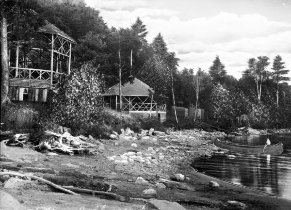 View of log pavilions along a shoreline at Cobbossee Colony (Camp?). The structures are surrounded by trees and a boy paddles a canoe near the sand on the beach.