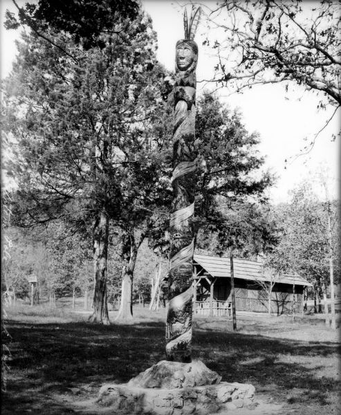 A totem pole stands in front of a wooden building in a wooded area. The pole features carvings of nature scenes, animals, and a Native American head wearing feathers.