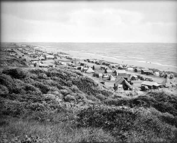 Tents line a beach along the Atlantic Ocean.  Foliage covers the foreground and several people and vehicles can be seen throughout the camp.
