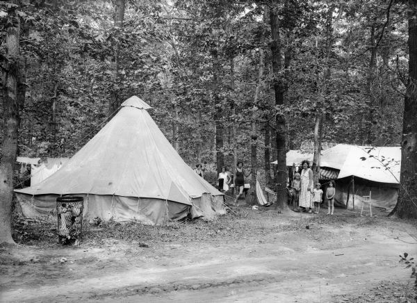 Several women and children wearing dresses or swimming suits gather around two tents set up in a wooded area.  A dirt road passes through the foreground.
