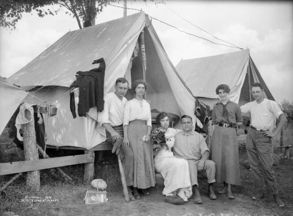 Three couples posing for a group portrait in front of tents at Mt. Tom Camp. The woman sitting at center is holding a bouquet of flowers, and clothing is hanging up to dry on a line at left.