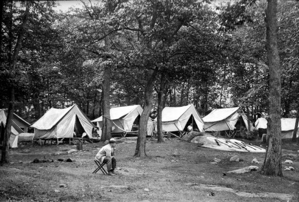 A row of tents at a Y.M.C.A. camp stands in a clearing in a wooded area.  A man sits on a chair in the foreground and "Y.M.C.A." is written on a surface off to the right.