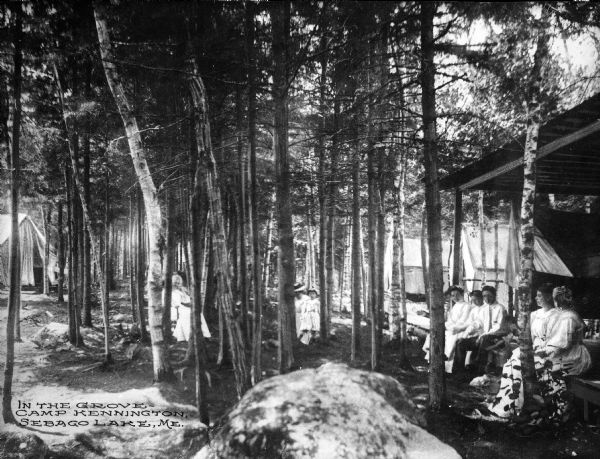Campers relax in front of their tents in a wooded grove at Camp Kennington. The women and children are wearing light-colored dresses, and the man is wearing dress pants, a shirt, and a necktie.