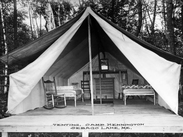 View of a the interior of a tent standing on a wooden platform at Camp Kennington. The tent flaps are open, showing two beds, a dresser, and a chair.