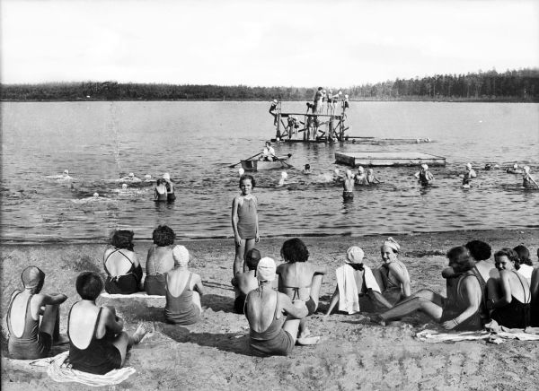 Girls and women sit along the Lutherland Beach at Camp Nawakwa.  Several others swim or stand on a wooden structure in the water.