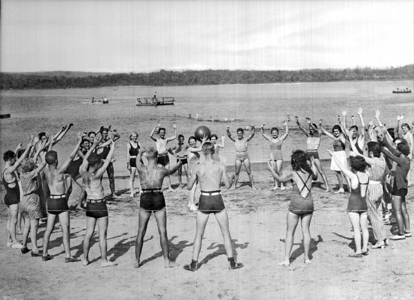 A group of people wearing swimming suits toss a beach ball on the sand at Beaver Brook. There are several swimmers, a dock, and several boats in the background.