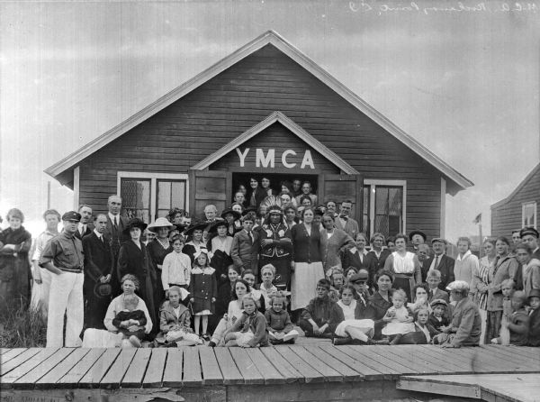 A group of people poses for a portrait in front of a Y.M.C.A. building.  A man dressed as a Native American chief stands at the center of the image and a wooden boardwalk passes through the foreground.