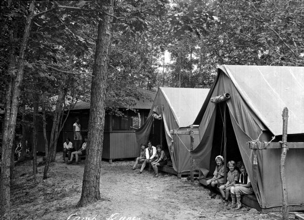 Two tents and two cabins stand in a clearing surrounded by trees.  Adults and children sit and stand in the doorways of the dwellings.