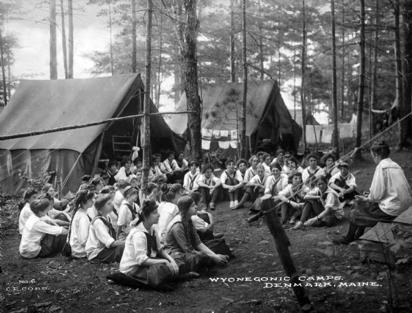 A group of uniformed female campers sits on the ground in front of their tents at Wyonegonic Camp while listening to their counselor.  Clothing hangs on lines strung between trees in the background.