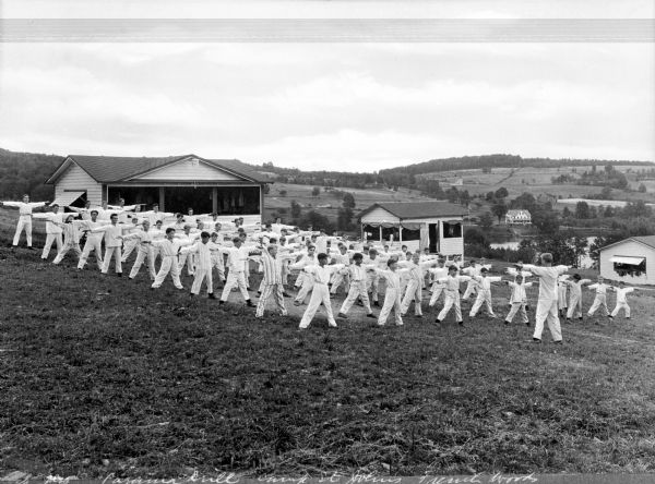 A group of boys at Camp St. John performs an exercise drill while wearing pajamas.  The group is gathered on a hilltop in front of cabins and farmhouses. Other dwellings are visible in the distance.