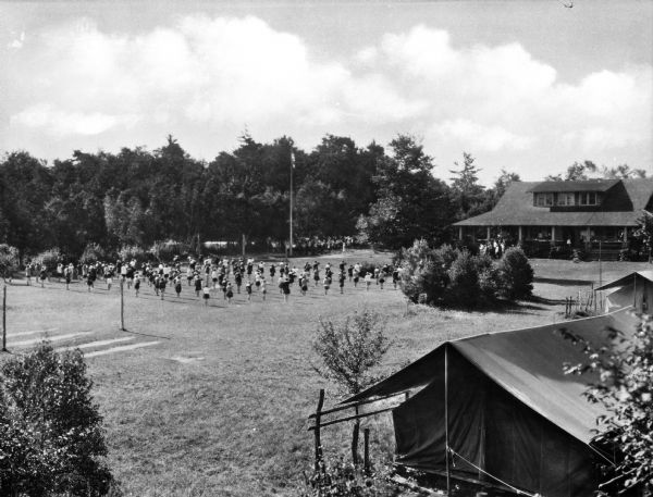 Elevated view of tents and girls exercising in formations at Pine Tree Camp. Trees and a flag pole stand to the left of the group and several tents stand in the foreground.