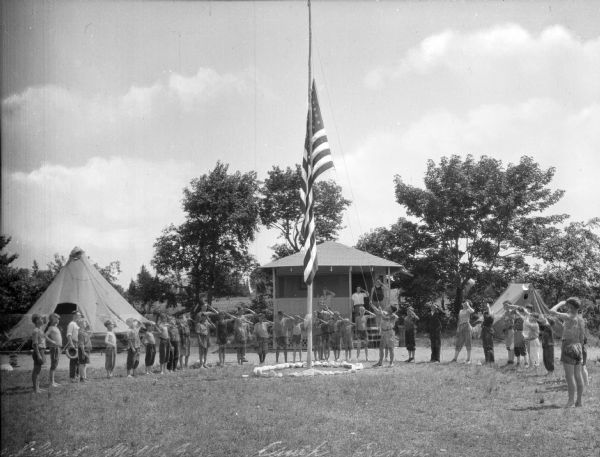 A group of boys and young men salutes while gathered in a semi-circle around a pole during the lowering of the American flag at Camp Gregory.  There are two tents and a cabin in the background, with a few people saluting from the porch of the cabin. Trees surround the camp site.