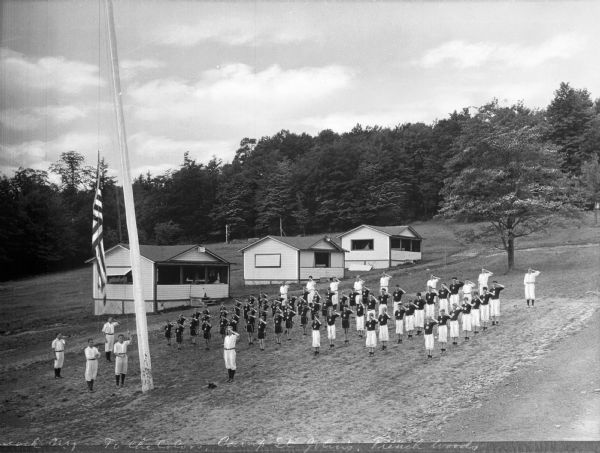 A group of uniformed campers stands in formation at Camp St. John while watching the raising of the American flag.  Three cabins stand to the left and trees are in the background.