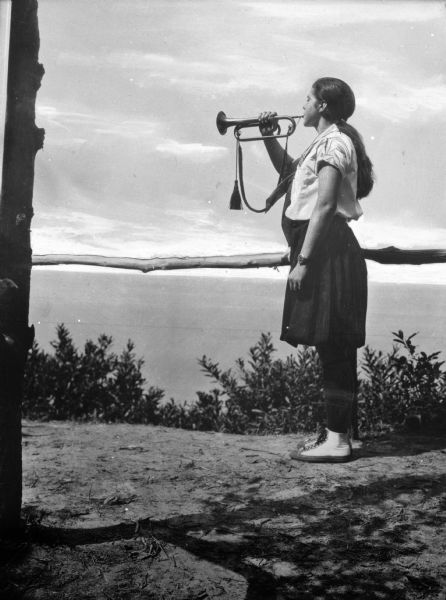 A girl wearing what appears to be a camp uniform-style dress plays a bugle on a hilltop.