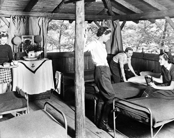 Four female campers stand and sit around a Y.W.C.A. camp cabin furnished with several beds and a dressing table.