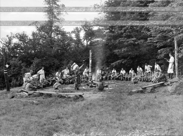 Campers watch Native American ceremonies while seated around the council ring at Camp St. John. The group of boys and young men gather around a fire pit, a totem pole, and a man in Native American dress. Trees surround the council ring.