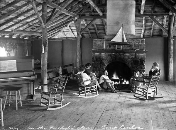 Three girls sit in rocking chairs and on the ground in front of a stone fireplace at Camp Lenoloc.  A model sailboat sits on the fireplace mantel and a piano and stool stand off to the left.