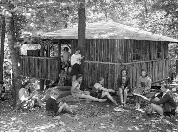 Girls gather around the entrance to the log Arts and Crafts cabin at Camp Lenoloc.  The cabin is elevated and surrounded by trees.
