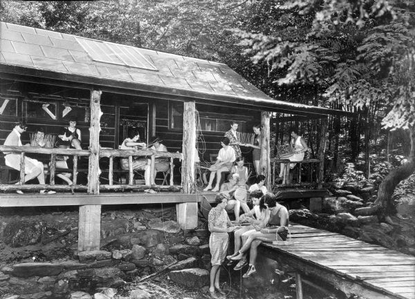 A group of campers sits on the porch of a cabin while making baskets at Camp Sherwood. Several others sit on a dock or wooden walkway leading from the cabin's porch.