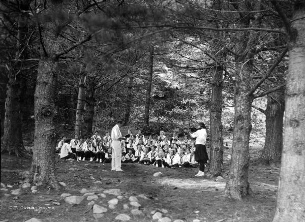 Uniformed campers sit on the ground while listening to a violin recital in the woods at Wyonegonic Camp.