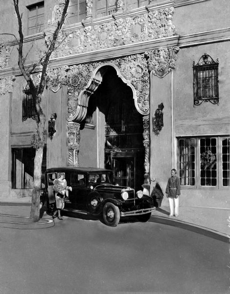 A Native American doorman stands at the street entrance of the Westward-Ho Hotel. View features an archway entrance, an automobile, and a woman standing to the side of the automobile.