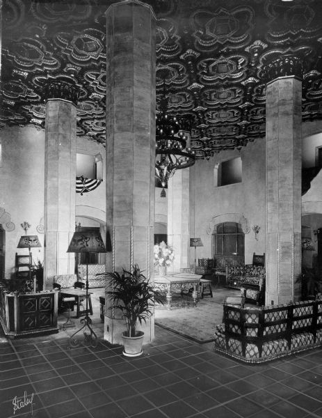 View of the lobby of the Westward-Ho Hotel. View features a decorative carved ceiling, furniture, columns, lamps, plants, tile floor, and a chandelier.