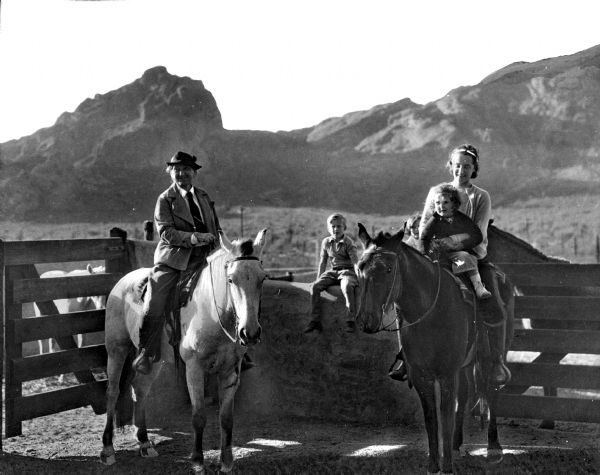 Hotel guests at Camelback Inn sit on horses near a fence in front of mountains.  A man sits on the horse to the left and a woman holds a child on the horse beside him while a boy looks on from the fence behind them.