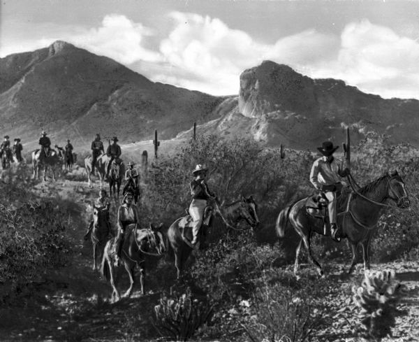 Guests at the Cambelback Inn ride horses along a mountain pass.  The trail is surrounded by foliage and cacti.