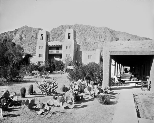 A covered patio leads to the Jokake Inn.  The building features two towers with a balcony between them, and the pavilion features outdoor furniture and seating. Cacti grow in the foreground and mountains stand in the background.