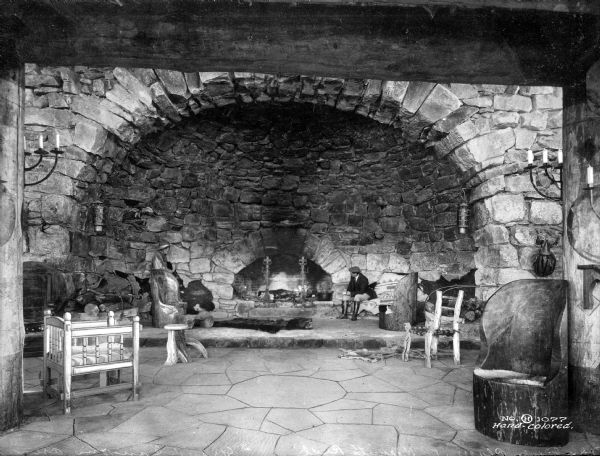 A man sits beside the stone fireplace at Hermit's Rest.  The build features a ceiling arch, chairs, and stone flooring.
