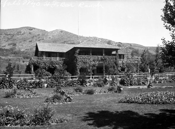 View of H.F. Bar Ranch House, a two-story ivy-covered log structure with a porch and several balconies.  A fence stands between the house and a lawn area with plants and a fountain.