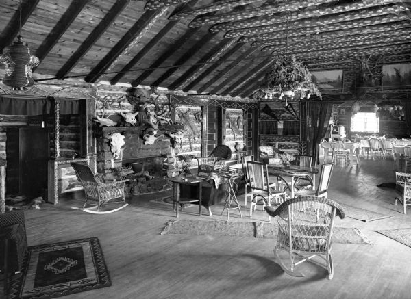 View of the rustic interior of a lodge. The building features a wood floor, angled wood ceiling, rugs, chandeliers, chairs and furniture and taxidermied animal heads and paintings hang on the walls. Tables and chairs can be seen off to the right in the next room.
