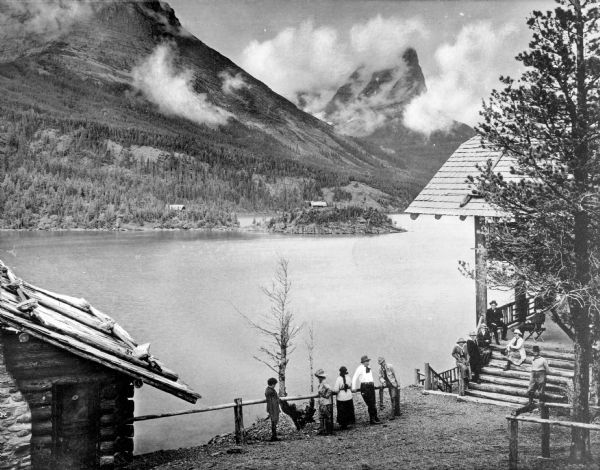 View of Going-to-the-Sun Chalets at Glacier National Park. View features rustic chalets overlooking the river and hills. Several people stand by the river while others sit and stand on the stairs of the chalet.
