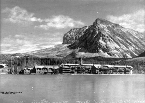 View of the Glacier Park Hotel from across Lake McDermott. View features the hotel surrounded by forest with a mountain in the background.