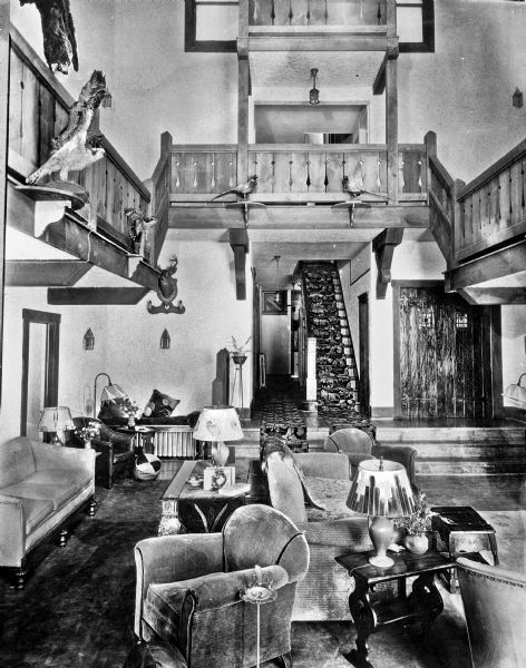 View of the lobby of the rustic Hunting Lodge at the Nahanock Country Club. View features a wood balcony overhead, a staircase, animal decorations, a doorway, and seating and furniture.