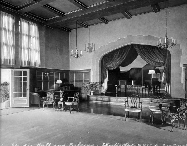 View of the Y.W.C.A. (210 E. 77th St.) studio and small stage. View features the stage on the right with curtains drawn, two chandeliers hanging in front of the stage, a doorway and windows to the left, and chairs and furniture arranged throughout the studio.