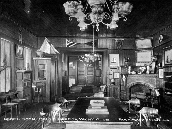 View of the Model Room at the Belle Harbor Yacht Club. View features a table and chairs, a fireplace on the right, chairs and furniture, several chandeliers, and a set of doors in the background. The room is decorated with flags, artwork, and a model sailboat.