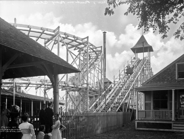 View of an amusement park featuring a roller coaster, a dwelling with a porch on the right, a pavilion, and a fence. Several people stand along the fence.