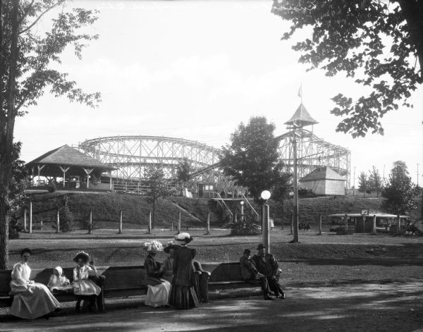 People gather around a bench in front of Casino Amusement Park.  A roller coaster, pavilion, and ticket booth stand behind them.
