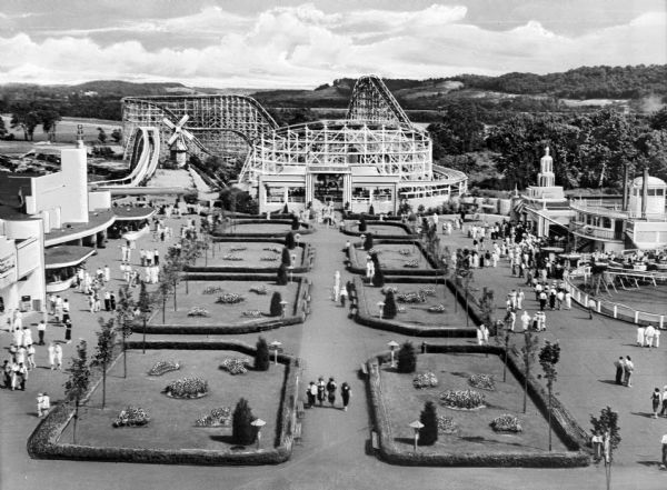 View of the Coney Island Amusement Park featuring a large garden and lawn, a roller coaster, a windmill, several buildings and amusements, and crowds of people.