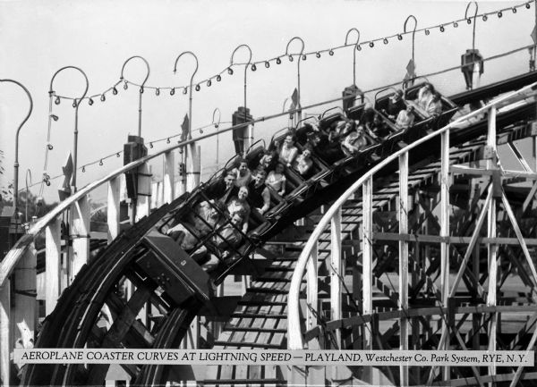 People ride a ten car roller coaster at PLAYLAND park.  The roller coaster has lights on one side and railings on either side.